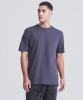 A man stands against a plain light grey background, wearing the AWDis 180 T dark grey short-sleeve t-shirt by AWDis and dark jeans. He looks slightly to his left with a neutral expression.