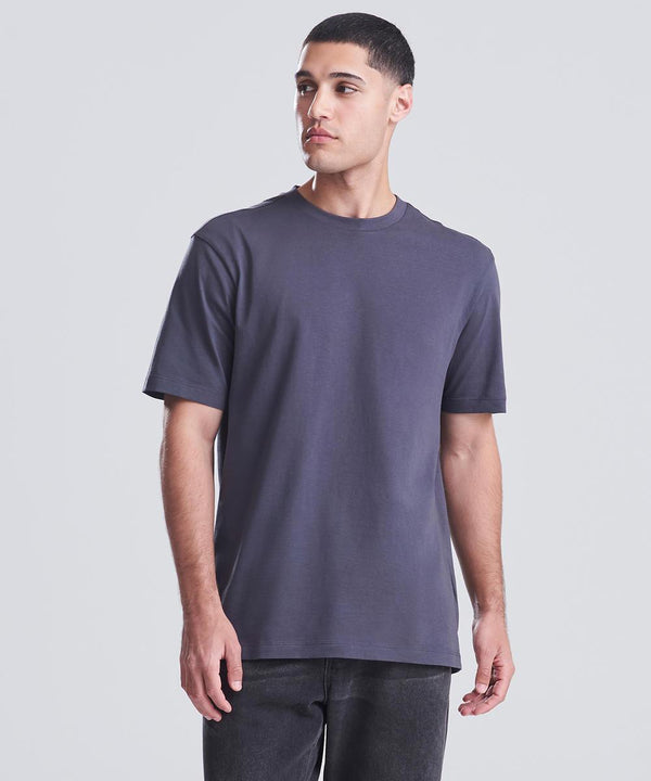 A man stands against a plain light grey background, wearing the AWDis 180 T dark grey short-sleeve t-shirt by AWDis and dark jeans. He looks slightly to his left with a neutral expression.