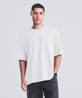 A person with short dark hair stands against a plain light background, wearing the AWDis 240 T by AWDis—a loose white t-shirt paired with dark trousers, one hand in their pocket and a neutral expression.