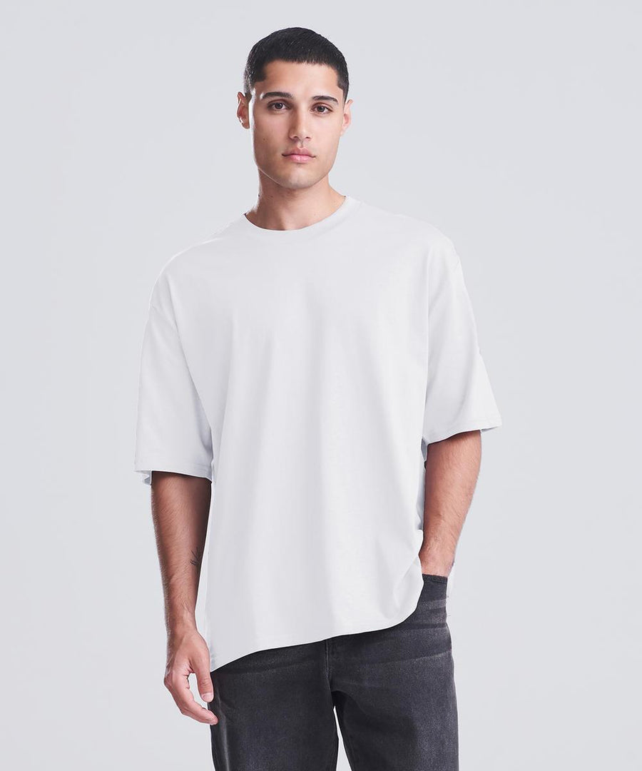 A person with short dark hair stands against a plain light background, wearing the AWDis 240 T by AWDis—a loose white t-shirt paired with dark trousers, one hand in their pocket and a neutral expression.