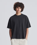 A young person with curly hair wears the AWDis 240 boxy cropped T in black and light-coloured trousers, standing against a plain light grey background with hands in pockets.