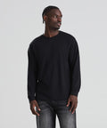 A man wearing an AWDis 180 long sleeve T in black and dark jeans stands against a light grey background, looking slightly to his left with his hands by his sides.