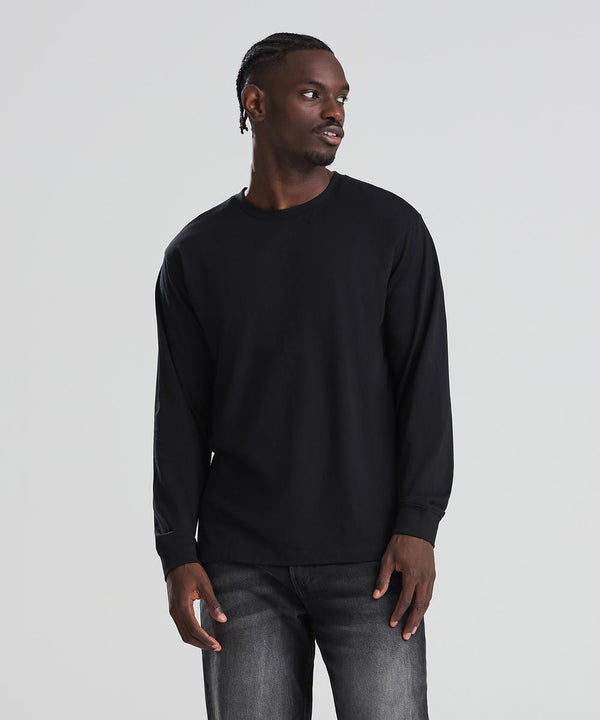 A man wearing an AWDis 180 long sleeve T in black and dark jeans stands against a light grey background, looking slightly to his left with his hands by his sides.