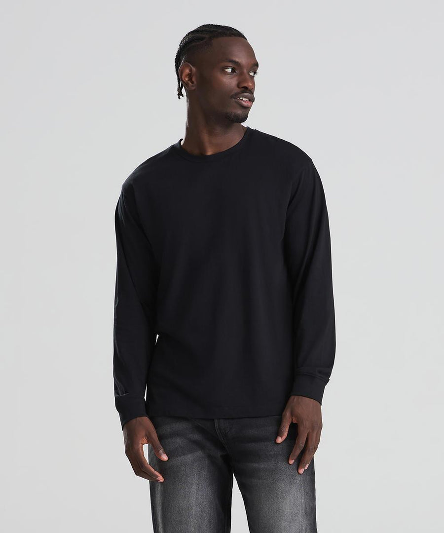 A man wearing an AWDis 180 long sleeve T in black and dark jeans stands against a light grey background, looking slightly to his left with his hands by his sides.
