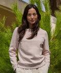 A woman with long dark hair stands outdoors in front of green foliage, wearing the B&C Queen crew neck sweatshirt by B&C Collection and light-coloured trousers. She has her hands in her pockets and looks at the camera neutrally.