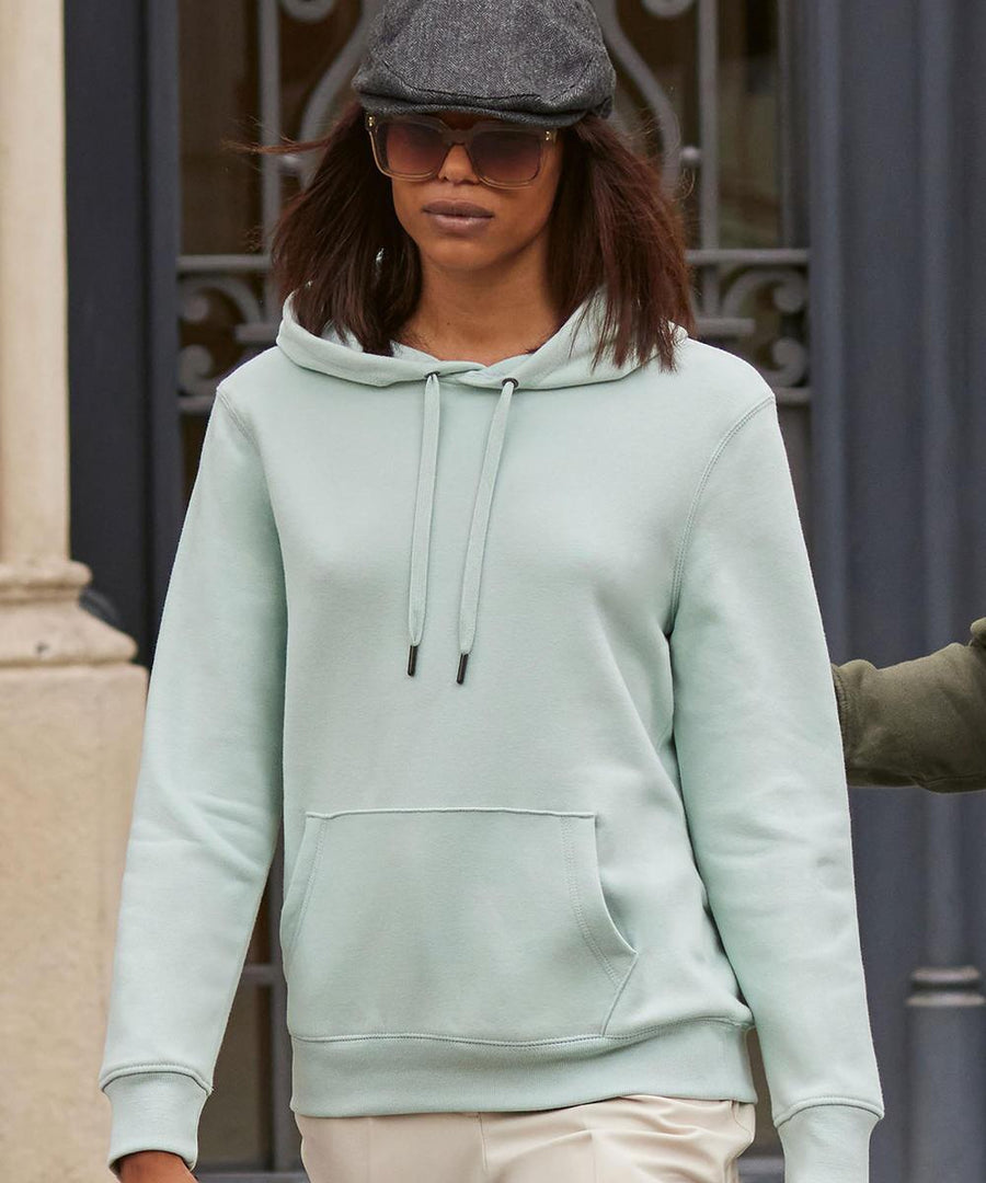 A person wearing a B&C Queen hooded light blue hoodie from B&C Collection, light-coloured trousers, dark sunglasses, and a grey flat cap stands outside in front of a decorative iron gate.