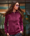 A woman wearing the B&C Collection B&C Queen zipped hoodie in maroon and purple-tinted sunglasses stands outdoors with her hands in her pockets, looking to the right, with a glass building façade in the background.