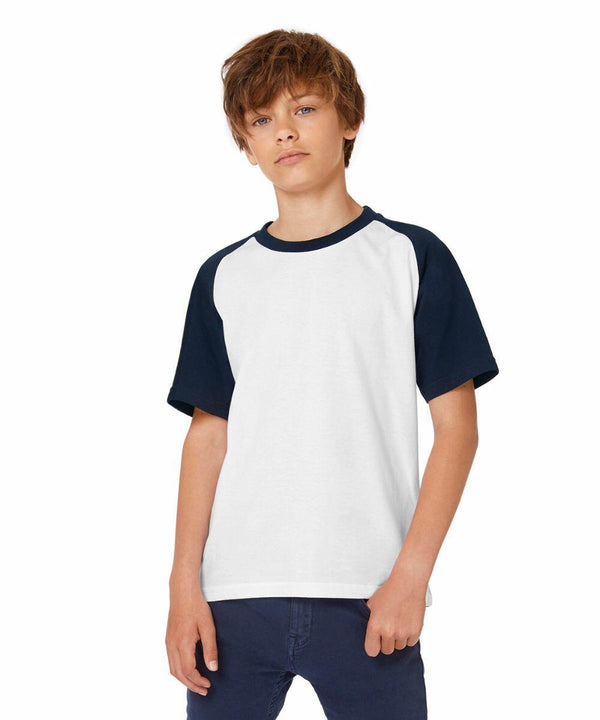 A young boy with shaggy, brown hair stands against a plain white background. He is wearing a white B111B T-shirt with dark blue sleeves and matching dark blue pants by B&C Collection. He has a neutral expression and his hands are relaxed at his sides.