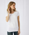 A person with long, light brown hair is standing against a plain white background. They are wearing a simple white B123F short-sleeve T-shirt by B&C Collection and dark jeans, looking to their left with their right hand gently touching their neck.