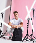 A young man dressed in a white t-shirt, light-colored pants, and sneakers squats in front of a pink background. He has a black Bagbase B125L backpack and is surrounded by tripods and bright neon lights forming an 'X' shape.