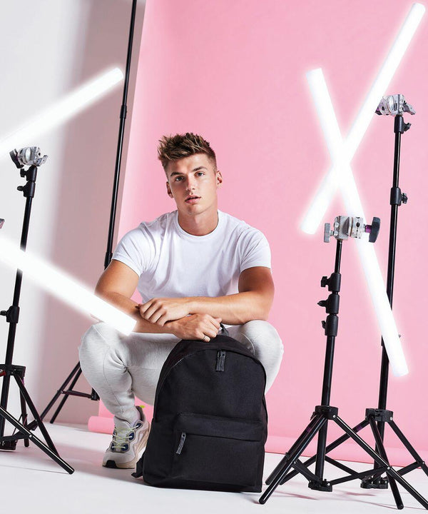 A young man dressed in a white t-shirt, light-colored pants, and sneakers squats in front of a pink background. He has a black Bagbase B125L backpack and is surrounded by tripods and bright neon lights forming an 'X' shape.