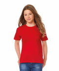 A young person with long, straight hair is wearing a plain red B150B by B&C Collection and blue jeans. They are standing against a white background and looking directly at the camera.