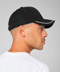 A person in profile wearing a Beechfield Match day cap in black with white trim and a white T-shirt stands against a plain grey background.