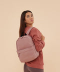 A woman with long brown hair stands before a beige background, looking over her shoulder. She wears a dusty pink sweatshirt and leggings, and carries the Bagbase Premium Recycled Mini Rucksack in light pink.