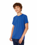 A young boy with short messy hair is standing against a plain white background. He is wearing a bright blue B190B T-shirt from B&C Collection and dark blue pants, with his right hand resting at his side and his left hand partially in his pocket. He has a slight smile on his face.