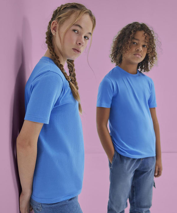 Two kids wear B&C Collection's B&C #E150 /kids blue T-shirts and jeans, posing against a pink background—one leans on the wall with arms at sides, the other stands with hands in pockets; both face the camera.