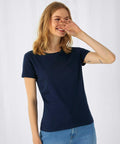 A woman with shoulder-length blonde hair is smiling and covering her mouth with her right hand. She is wearing a navy blue B210F short-sleeve shirt from B&C Collection and light blue jeans. The background is plain white. She has a small tattoo on her left forearm.