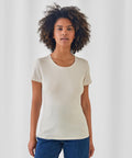 A woman with curly hair faces the camera, wearing a short-sleeved off-white B&C #Inspire E150 /women t-shirt from B&C Collection and blue jeans, set against a plain light background.