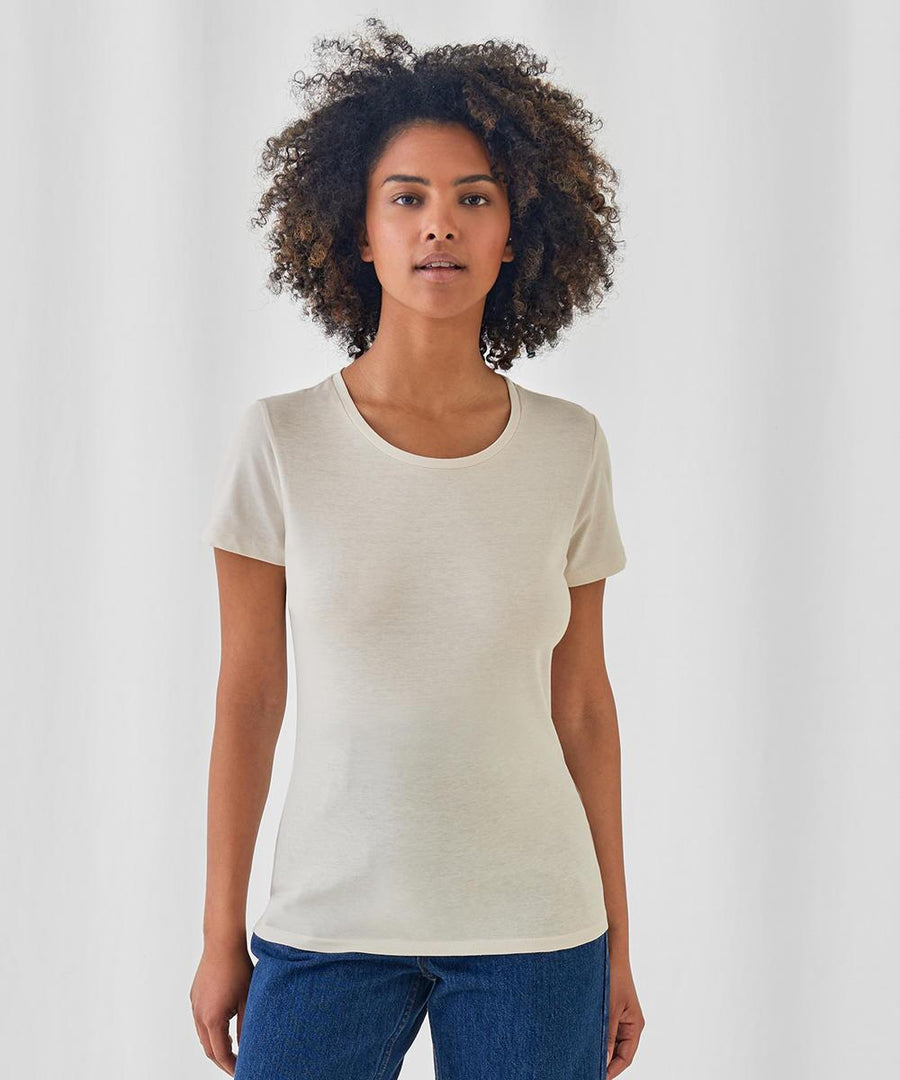A woman with curly hair faces the camera, wearing a short-sleeved off-white B&C #Inspire E150 /women t-shirt from B&C Collection and blue jeans, set against a plain light background.