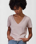 A woman with curly hair wears the B&C Collection B&C #E150 V /women light pink V-neck short-sleeved T-shirt and light blue jeans, standing with her hands in her pockets and looking down against a plain background.