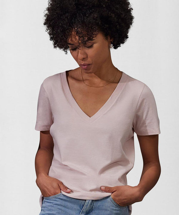 A woman with curly hair wears the B&C Collection B&C #E150 V /women light pink V-neck short-sleeved T-shirt and light blue jeans, standing with her hands in her pockets and looking down against a plain background.