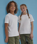 Two children wearing B&C Collection's B&C #E190 /kids plain white t-shirts and green cargo trousers stand against a blue wall. One has curly hair, the other two braided plaits; both face the camera with slight smiles.