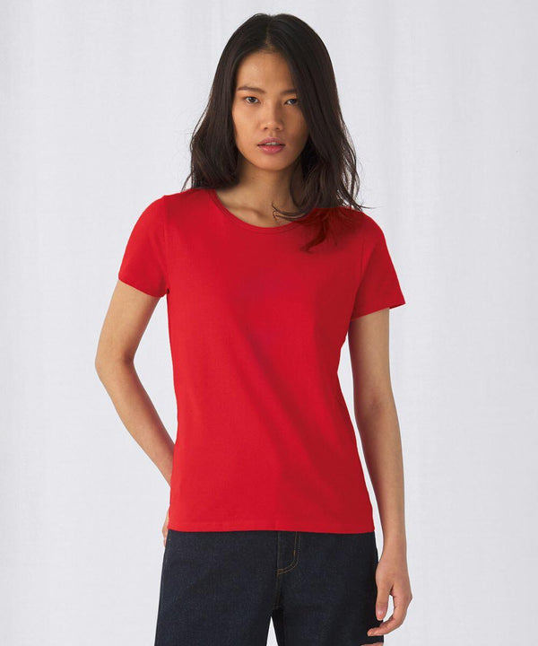 A person with long, dark hair is wearing a plain red short-sleeve B220F t-shirt from B&C Collection and dark jeans. They are standing against a white background, looking at the camera.