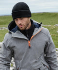 A man with a beard is wearing a gray jacket with an orange zipper and a Beechfield B243R. He is looking down, with an outdoor grassy landscape and a cloudy sky in the background.