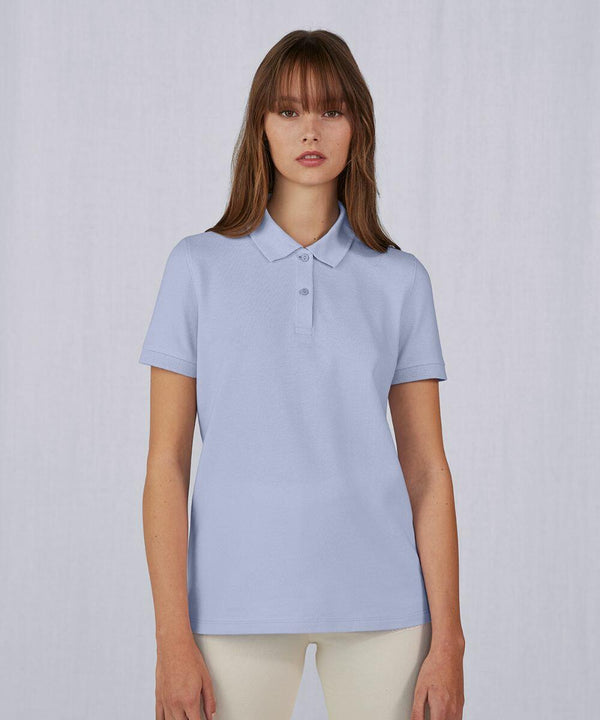 A young woman with straight brown hair and bangs is wearing a B261F polo shirt from B&C Collection. She is standing against a plain, light-colored background. The shirt has a classic collar and three buttons down the front.