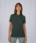 A person with long brown hair is standing against a plain white background. They are wearing a dark green short-sleeved B263F by B&C Collection and light gray pants. Their expression is neutral, with eyes looking slightly downwards.