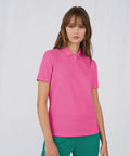 A woman with long brown hair wearing a bright pink B265F polo shirt from B&C Collection and green pants stands against a plain white background. She has a neutral expression and her arms are relaxed by her sides.