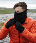 A person wearing a bright orange jacket and black gloves is outdoors in a rocky landscape. They are pulling up a black Beechfield B280R fleece neck gaiter to cover their face, with a grassy background visible. The sky is overcast.