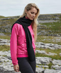 A woman with long blonde hair stands outdoors on a rocky terrain. She is wearing a bright pink jacket, black scarf, and dark pants. She appears to be looking down pensively. The background features grass, rocks, and a cloudy sky, accented by the Beechfield B293R she's bundled in.