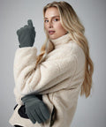 A person with long blonde hair wears a cream-colored Beechfield B298R fleece jacket and dark gray gloves. They are gazing at the camera with a slight smile, holding their right hand up near their face while the left hand is positioned by their side. The background is plain gray.