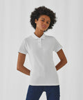 A person with short curly hair is standing against a plain white background, wearing a B&C Collection B306F polo shirt and blue jeans. The individual's hands are positioned behind their back, and they are looking directly at the camera with a neutral expression.