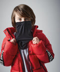 A child with brown hair wearing a bright red puffer jacket with lettering on the sleeves pulls up a black Beechfield B320B over their nose, against a plain gray background. The child looks directly at the camera.