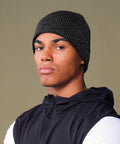 A person models the Beechfield Streetlite reflective beanie in black and white, paired with a black jacket with white sleeves, standing against a light olive background and looking at the camera with a neutral expression.