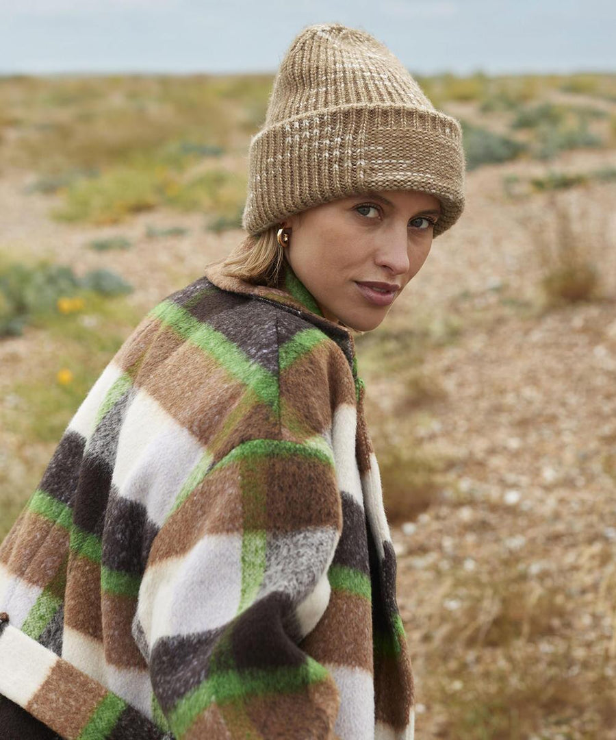 Wearing a Beechfield Chunky Knit Reverse Patch Beanie, a person looks over their shoulder outdoors on a rocky, grassy landscape, styled with gold hoop earrings and a brown, green, and white checked coat.