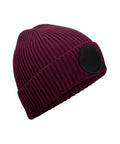 A maroon ribbed knit beanie with a folded brim and a prominent black circular patch on the side, shown on a white background.