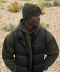 Someone outdoors on a rocky surface, facing sideways, wears a green Circular fashion patch beanie by Beechfield, paired with a green ribbed hoodie and black puffer gilet.