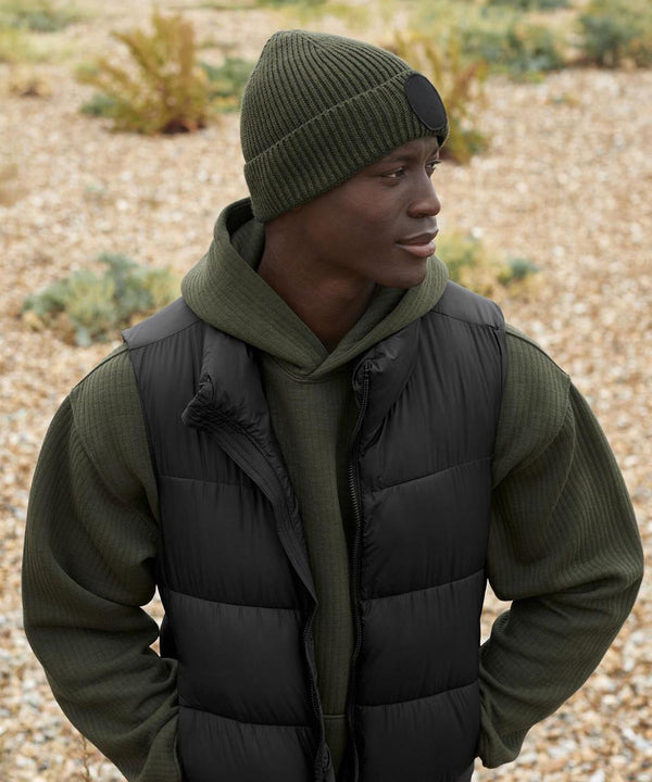 Someone outdoors on a rocky surface, facing sideways, wears a green Circular fashion patch beanie by Beechfield, paired with a green ribbed hoodie and black puffer gilet.