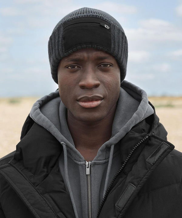 Wearing a Beechfield Zip patch beanie in black, a grey hoodie, and a black jacket, the person stands outdoors on a cloudy day with a neutral expression; the background shows an out-of-focus sandy or gravelly area.