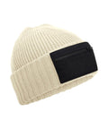 The Beanies is a beige knitted beanie with a wide folded brim and a rectangular black patch featuring a front zip pocket.
