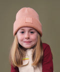A young girl with straight, light brown hair wears the Beechfield Kids Deep Cuffed Tonal Patch Beanie in peach and a beige waistcoat over a maroon long-sleeve shirt, posing before a plain olive-green backdrop.