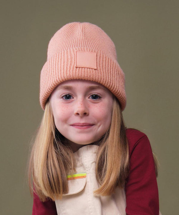 A young girl with straight, light brown hair wears the Beechfield Kids Deep Cuffed Tonal Patch Beanie in peach and a beige waistcoat over a maroon long-sleeve shirt, posing before a plain olive-green backdrop.