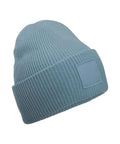 A beanie hat in light blue features ribbed knit fabric, a cuffed brim, and a rectangular front patch, displayed on a plain white background.