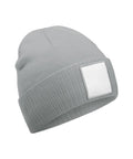 A grey knitted beanie hat with a plain white rectangular patch on the front turn-up is shown against a white background.