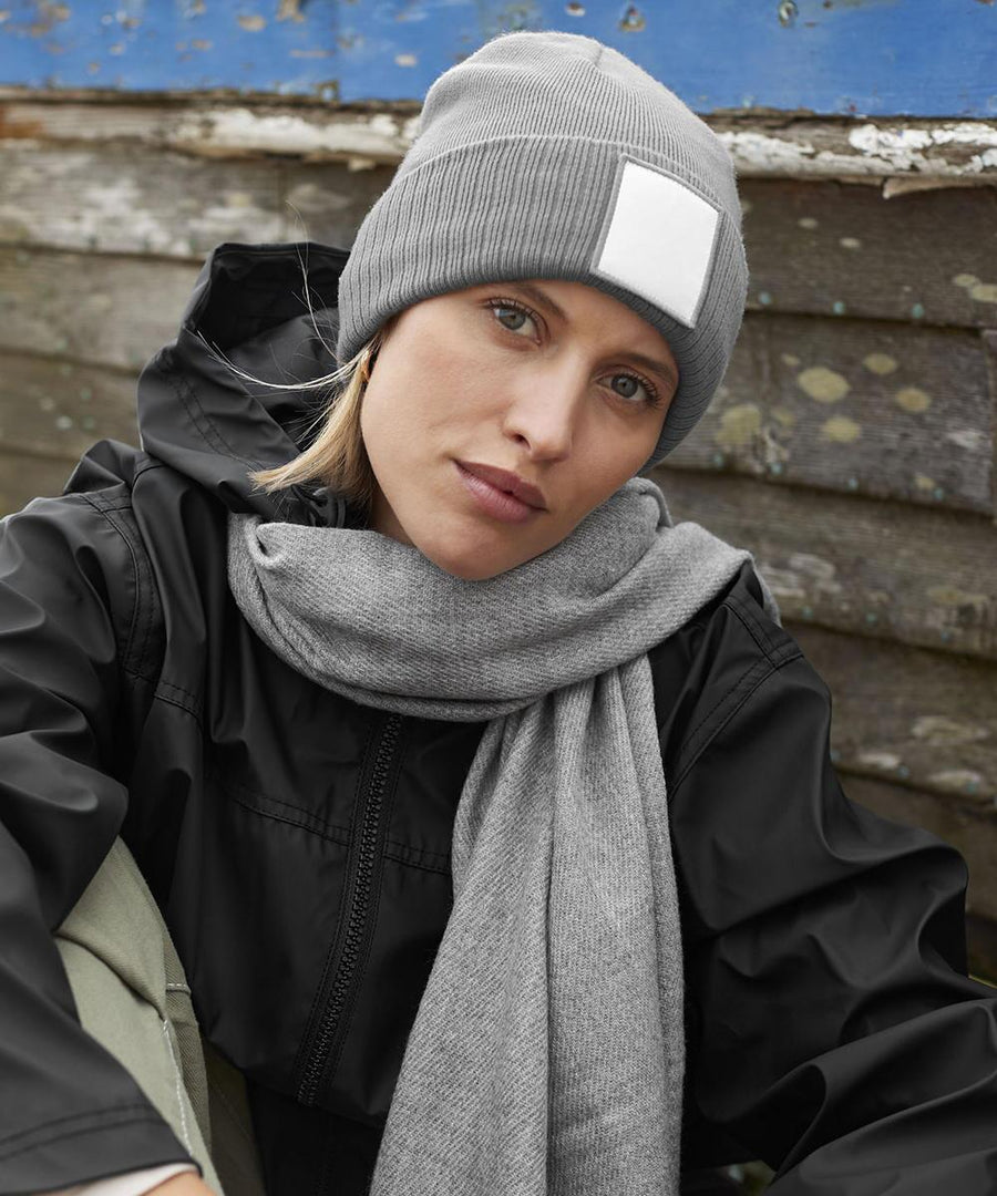 A person wearing a grey Beechfield appliqué patch beanie, a grey scarf, and a black jacket sits in front of a weathered wooden wall with blue paint at the top.