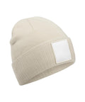 A beige knitted beanie hat with a wide folded brim and a blank white rectangular patch on the front, displayed against a plain white background.