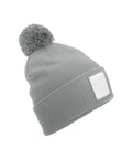 A grey knitted beanie hat with a pom-pom on top and a blank white rectangular patch on the front, displayed against a white background.